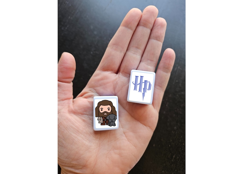 May include: Two white square tiles with images from the Harry Potter universe. One tile features the Harry Potter logo with a lightning bolt in purple. The other tile features a cartoon image of  Rubeus Hagrid with his pet dog Fang.