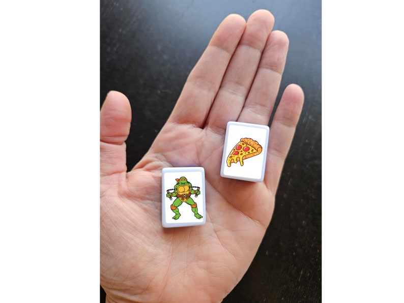 May include: Two white square tiles with images. One tile has a cartoon image of a Teenage Mutant Ninja Turtle. The other tile has a cartoon image of a slice of pizza.