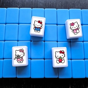 May include: Four white tiles with Hello Kitty images on a blue background. The tiles feature Hello Kitty in different outfits, including a blue dress, a pink dress, a pink dress with a camera, and a pink dress with a strawberry.