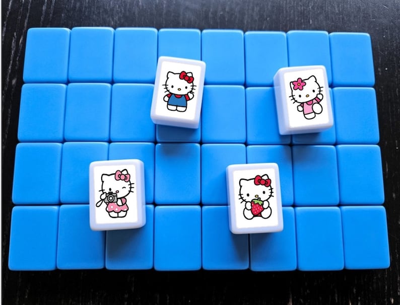 May include: Four white tiles with Hello Kitty images on a blue background. The tiles feature Hello Kitty in different outfits, including a blue dress, a pink dress, a pink dress with a camera, and a pink dress with a strawberry.
