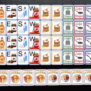 May include: A set of 16 white square tiles with colorful images and text. The tiles feature images of various breakfast foods, including waffles, crepes, pancakes, syrup, and whipped cream. The text on the tiles includes the words "Waffle Dragon", "Crepe Dragon", "Pancake Dragon", "N", "E", "S", and "W". There are also 8 smaller white square tiles with the word "JOKER" and images of breakfast foods.