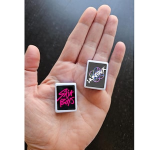May include: Two small, rectangular white objects with black faces are held in a hand. One has the text "SAJA BOYS" in pink, the other has "HUNTIX" with a colorful design. The objects appear to be small cards or tokens.