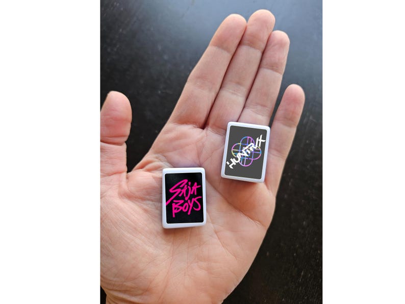 May include: Two small, rectangular white objects with black faces are held in a hand. One has the text "SAJA BOYS" in pink, the other has "HUNTIX" with a colorful design. The objects appear to be small cards or tokens.