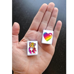Op de afbeelding: Twee witte vierkante tegels met kleurrijke afbeeldingen. E&eacute;n tegel heeft een regenboog hart, de andere tegel heeft een cartoon tijgerwelp.