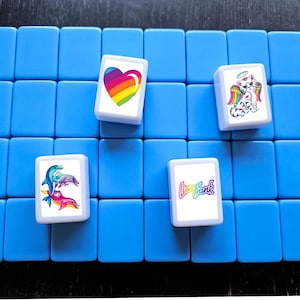 Op de afbeelding: Vier kleurrijke tegels met verschillende ontwerpen op een blauwe achtergrond. De tegels tonen een regenboog hart, een kat met engelenvleugels, een groep dolfijnen en de tekst "Good Frank".