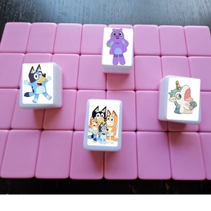 K&ouml;nnte beinhalten: Vier quadratische Fliesen mit Comicfiguren darauf. Die Fliesen sind wei&szlig; mit rosa R&auml;ndern und auf einer rosa Oberfl&auml;che angeordnet. Die Figuren sind ein blauer Hund, ein lila Monster, ein blaues Einhorn und eine Gruppe von drei blauen Hunden.