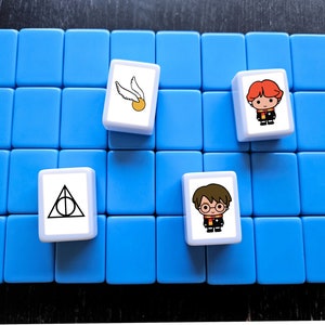 May include: A blue game board with four white tiles. Each tile has a different image from the Harry Potter universe. One tile has a golden snitch and a broom, another has the Deathly Hallows symbol, and the other two have cartoon images of Harry Potter characters.