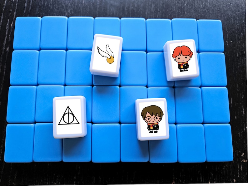 May include: A blue game board with four white tiles. Each tile has a different image from the Harry Potter universe. One tile has a golden snitch and a broom, another has the Deathly Hallows symbol, and the other two have cartoon images of Harry Potter characters.