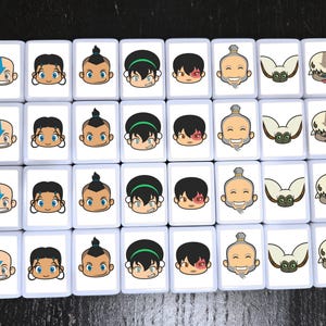 May include: A collection of white rectangular tiles, each featuring a cartoon character's face. The characters include a bald boy with an arrow tattoo, a girl with dark hair, a boy with a topknot, a girl with a green headband, a boy with a scar, an elderly man, a flying bison, and a lemur.