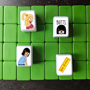 K&ouml;nnte beinhalten: Vier wei&szlig;e quadratische Kacheln mit Bildern von Figuren aus der Zeichentrickserie Bob's Burgers. Die Kacheln sind auf einer gr&uuml;nen Oberfl&auml;che angeordnet. Die Kacheln zeigen den Text "Bob's Burgers", "Butts" und Bilder der Figuren Tina und Louise Belcher.