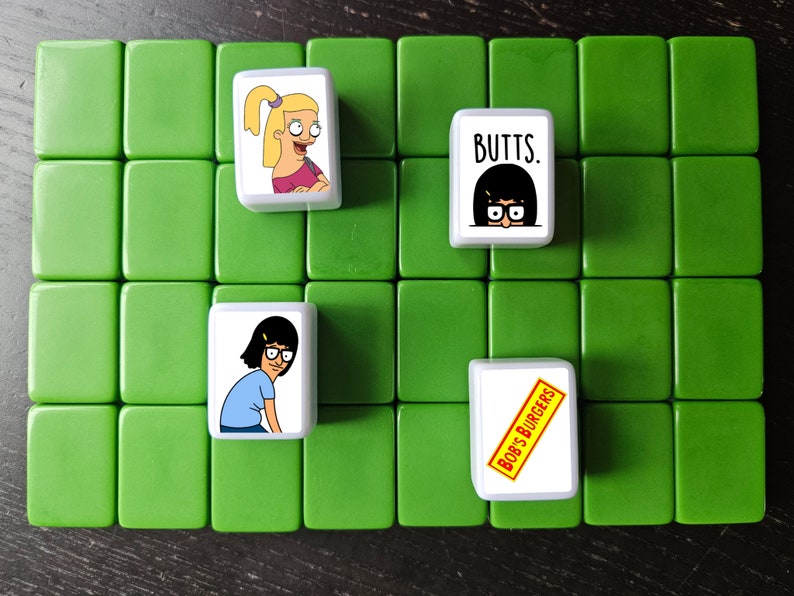 K&ouml;nnte beinhalten: Vier wei&szlig;e quadratische Kacheln mit Bildern von Figuren aus der Zeichentrickserie Bob's Burgers. Die Kacheln sind auf einer gr&uuml;nen Oberfl&auml;che angeordnet. Die Kacheln zeigen den Text "Bob's Burgers", "Butts" und Bilder der Figuren Tina und Louise Belcher.