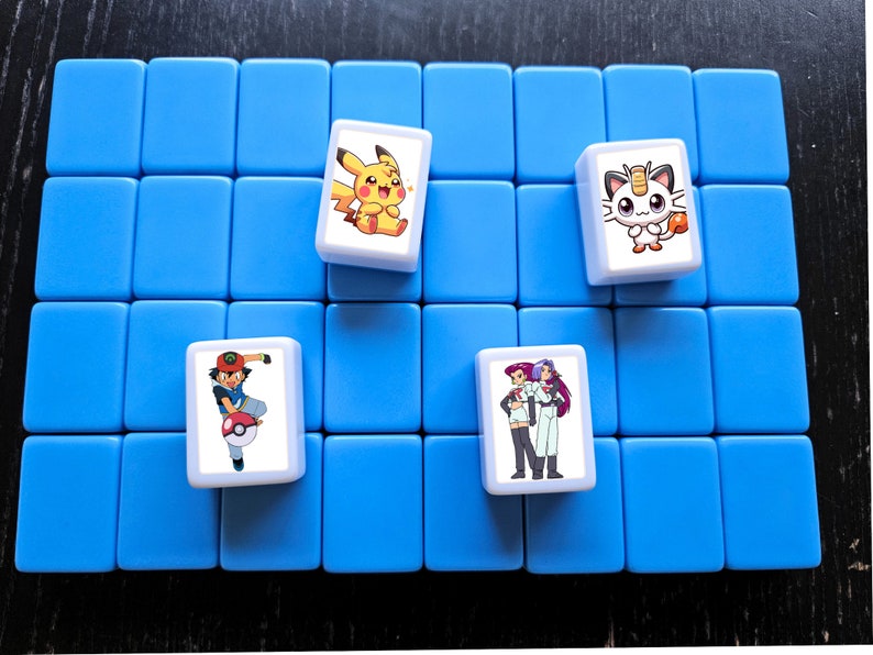 May include: A blue game board with four white tiles featuring Pokemon characters. The tiles show Pikachu, Eevee, Ash Ketchum, and  Brock and Misty.