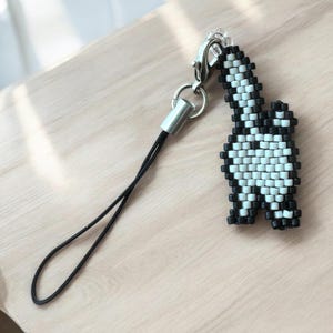 Puede incluir: Un dije de cuentas blanco y negro con forma de dinosaurio. El dije está sujeto a un cierre de metal plateado y un lazo de cordón negro. El dije está hecho de pequeñas cuentas cuadradas.