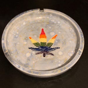 May include: A round ceramic ashtray with a blue and white speckled glaze. A rainbow marijuana leaf is painted in the centre of the ashtray.