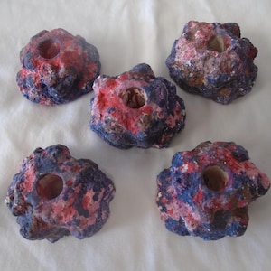 May include: Five decorative candle holders with a rock-like appearance. Each holder is painted with a combination of red, blue, and white colors. The holders have a hole in the center for a candle.
