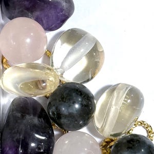 May include: A close-up of a collection of tumbled stones, including amethyst, rose quartz, clear quartz, and black obsidian. The stones are arranged in a cluster and are surrounded by a gold chain.