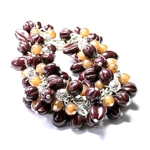 Puede incluir: Un brazalete de cuentas marrones y beige con una cadena plateada. Las cuentas tienen forma de pequeñas bellotas redondeadas.