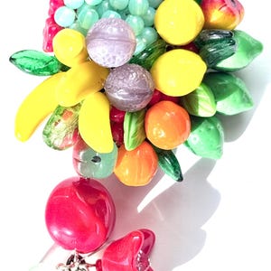 May include: A colourful fruit brooch made of glass beads. The brooch features a variety of fruits, including lemons, oranges, strawberries, and bananas. The fruits are arranged in a cluster and are surrounded by green leaves. The brooch is attached to a silver chain.