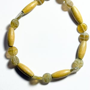 Puede incluir: Un brazalete de cuentas amarillas y verdes con un cierre dorado. El brazalete está hecho de cuentas de madera y vidrio.