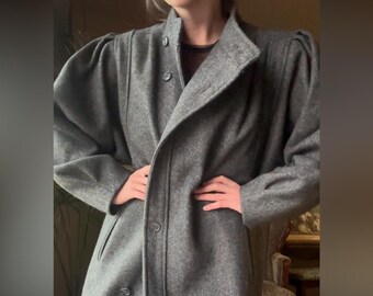 Puede incluir: Un abrigo de lana gris con cuello alto y cierre de botones. El abrigo presenta un diseño cruzado y hombros acolchados. El abrigo es de estilo de longitud media, adecuado para climas más fríos.