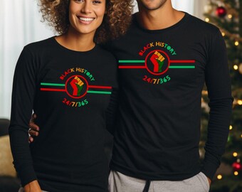 Puede incluir: Camiseta negra de manga larga con un diseño gráfico rojo, verde y negro. El gráfico presenta un puño levantado en un círculo con el texto "Black History 24/7/365".