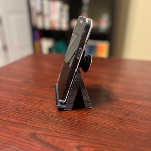 May include: A black phone stand with a phone propped up on it. The phone has a black case and a pop socket.