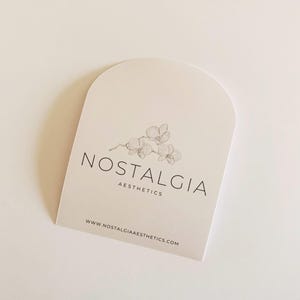 May include: A cream-colored, arched card featuring the text "NOSTALGIA AESTHETICS" in black, with a delicate orchid illustration. The website address www.nostalgiaaesthetics.com is printed at the bottom.