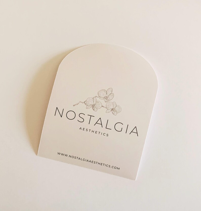 May include: A cream-colored, arched card featuring the text "NOSTALGIA AESTHETICS" in black, with a delicate orchid illustration. The website address www.nostalgiaaesthetics.com is printed at the bottom.