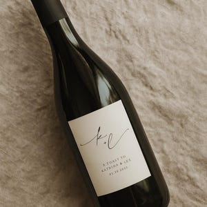 May include: A dark wine bottle with a white label featuring the initials "k + l" in elegant script. The label also includes the text "A TOAST TO KATRINA & LEX 01.10.2033". The bottle is set against a neutral, textured background.