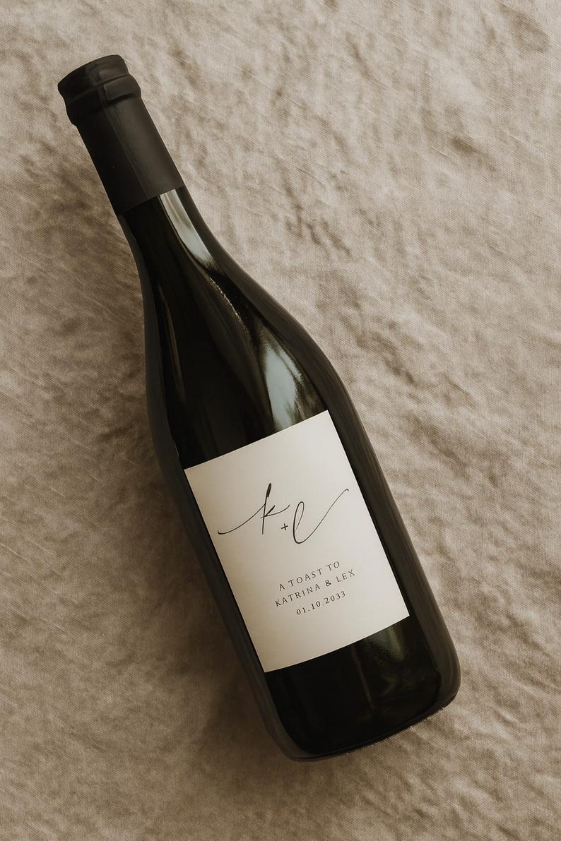 May include: A dark wine bottle with a white label featuring the initials "k + l" in elegant script. The label also includes the text "A TOAST TO KATRINA & LEX 01.10.2033". The bottle is set against a neutral, textured background.