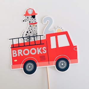 May include: A red fire engine cake topper with the name "BROOKS" printed on the side. A Dalmatian dog wearing a firefighter's hat sits on top. A number "2" and a sun are also included.