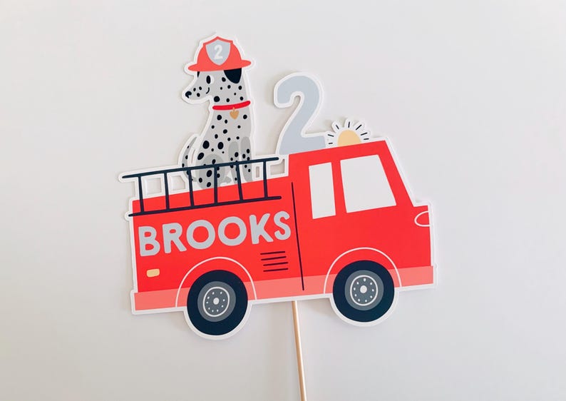May include: A red fire engine cake topper with the name "BROOKS" printed on the side. A Dalmatian dog wearing a firefighter's hat sits on top. A number "2" and a sun are also included.