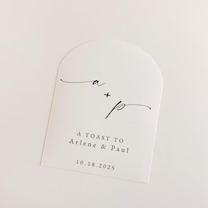 May include: White arched card with black script. The card features the initials "a + p" in elegant cursive, followed by the text "A TOAST TO Arlene & Paul" and the date "10.18.2025". The design is minimalist and suitable for wedding stationery.