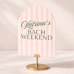 May include: A sign with a pink and white striped background and the text "Katrina's BACH WEEKEND" in green script. The sign is held by a gold-colored metal stand with a round base.