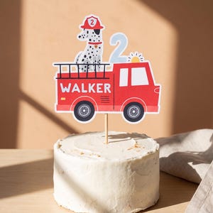 May include: A cake topper featuring a red fire truck with the name WALKER printed on the side, a dalmatian dog wearing a red firefighter helmet with the number 2, and a light blue number 2, all attached to a wooden stick inserted into a cake.