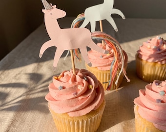 Può includere: Cupcakes con glassa rosa e piccole decorazioni a forma di stella, sormontati da sagome di unicorni in carta. Gli unicorni sono rosa e bianchi con corna glitterate argentate e code in filo multicolore fissate su stecchini di legno.