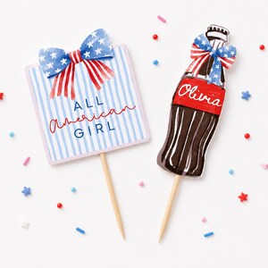 K&ouml;nnte beinhalten: Zwei patriotisch gestaltete Tortenaufs&auml;tze. Einer ist ein rechteckiges Schild mit blau-wei&szlig;en Streifen, einer Schleife und dem Text "ALL AMERICAN GIRL". Der andere ist eine braune Flasche mit einem roten Etikett mit der Aufschrift "Olivia" und einer Schleife.