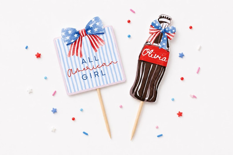 K&ouml;nnte beinhalten: Zwei patriotisch gestaltete Tortenaufs&auml;tze. Einer ist ein rechteckiges Schild mit blau-wei&szlig;en Streifen, einer Schleife und dem Text "ALL AMERICAN GIRL". Der andere ist eine braune Flasche mit einem roten Etikett mit der Aufschrift "Olivia" und einer Schleife.