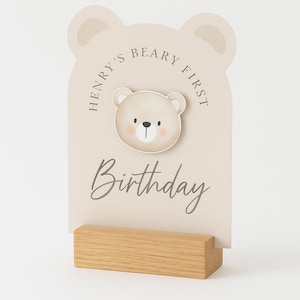 May include: A beige birthday sign with a bear illustration and the text "Henry's Beary First Birthday." The sign is mounted on a wooden base. The bear has a pink nose and is centered on the sign.