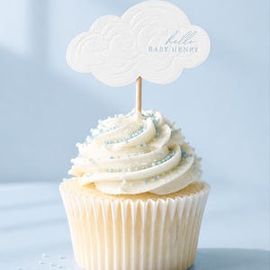 May include: A cupcake with white frosting and blue sprinkles, topped with a cloud-shaped pick. The pick has the text "hello BABY HENRY" in blue. The cupcake sits in a white paper liner against a light blue background.
