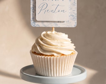 May include: A vanilla cupcake with white frosting sits on a light blue pedestal stand. A square topper features a blue and white toile pattern with a giraffe and hot air balloon, displaying the text BABY Preston in blue serif and script fonts.