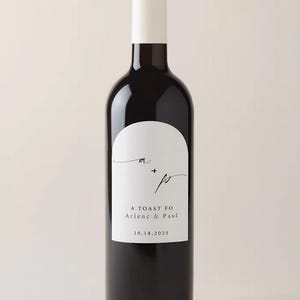 May include: A dark wine bottle with a white label featuring the initials "a" and "p" with a plus sign, and the text "A Toast Fo Arlene & Paul 10.18.2025". The bottle has a white top.