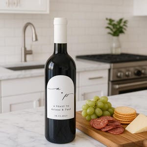 May include: A black wine bottle with a white label that reads "A Toast to Arlene & Paul" and the date 10.13.2023. The bottle is next to a wooden board with grapes, salami, crackers, and cheese. The kitchen has white cabinets and a stainless steel stove.