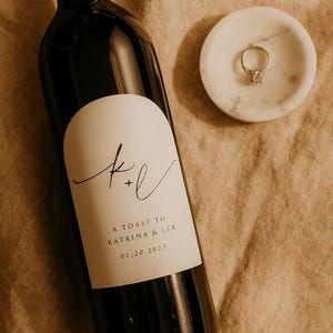 May include: A dark wine bottle with a white label featuring the initials "k+l" and the text "A TOAST TO KATRINA & LEX 01.20.2025". A marble ring dish holds a diamond ring. The items are on a tan surface.
