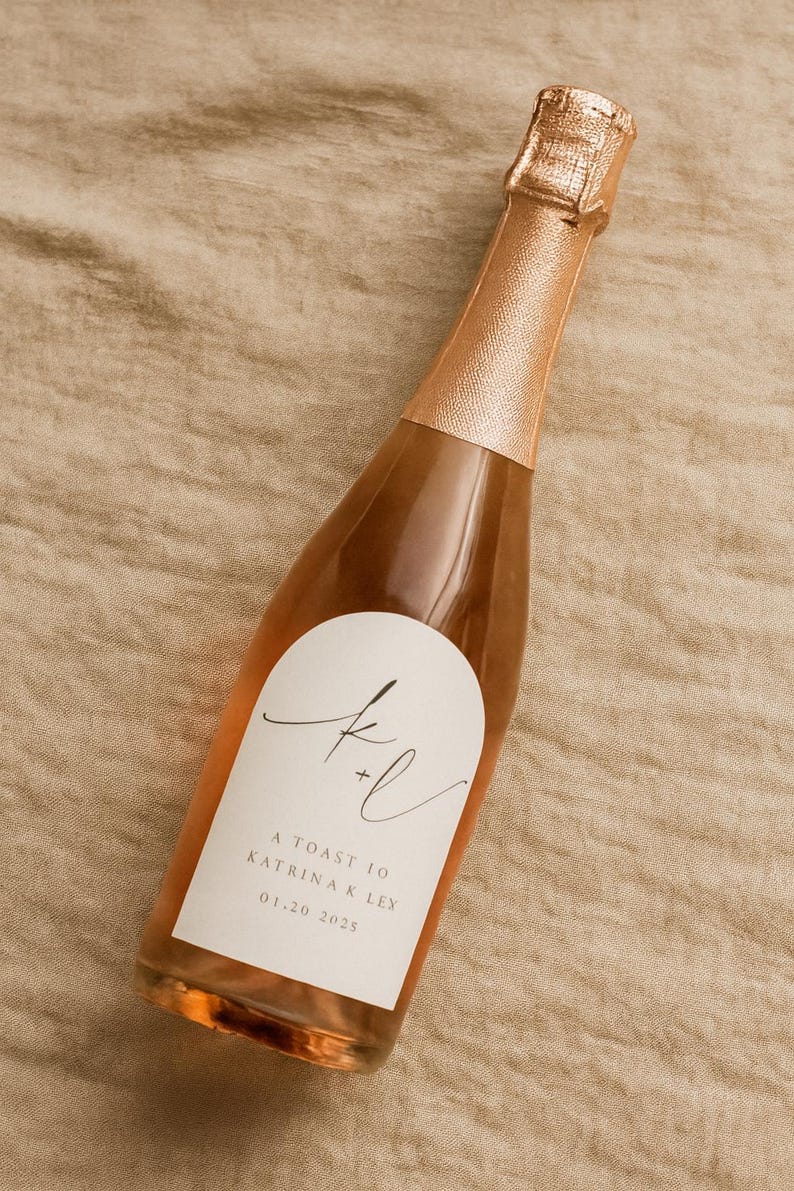 May include: A small bottle of sparkling wine with a rose gold foil top and label. The label has elegant script with the text "A Toast to Katrina K Ley 01.20.2025". The bottle is filled with a light pink liquid, suggesting a ros&eacute; or champagne.