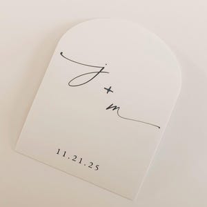 May include: A white, arched card with black calligraphy. The card features the initials "J + m" and the date "11.21.25" printed in a simple, elegant font. The design is minimalist and suitable for wedding or event announcements.