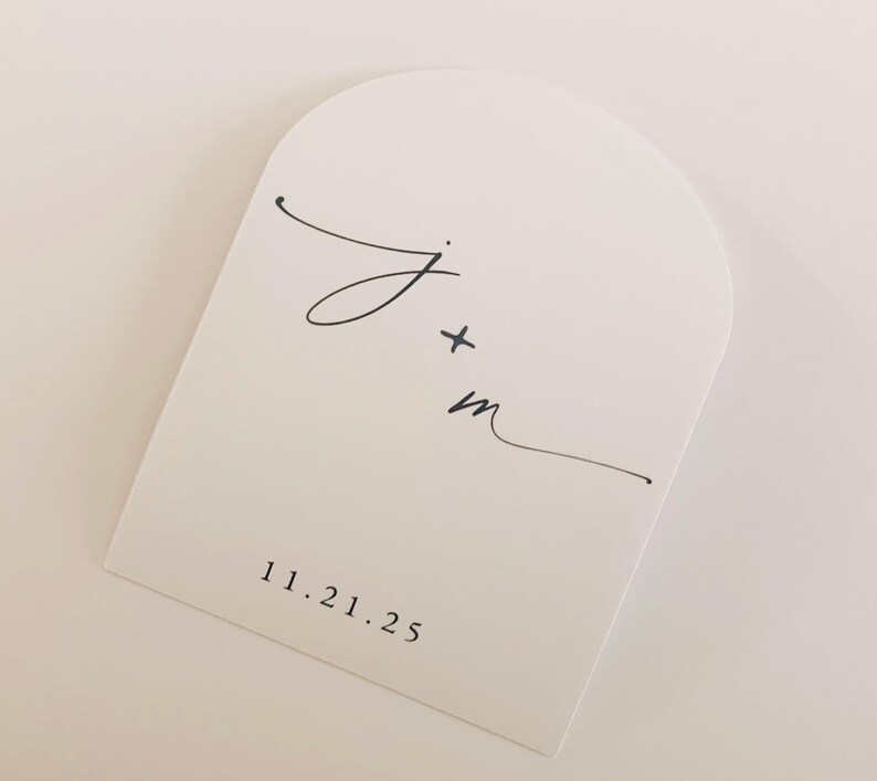 May include: A white, arched card with black calligraphy. The card features the initials "J + m" and the date "11.21.25" printed in a simple, elegant font. The design is minimalist and suitable for wedding or event announcements.