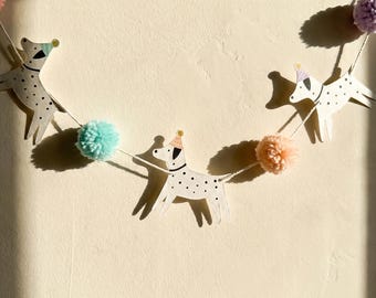 May include: A decorative garland featuring three white dog cutouts with black spots, each wearing a small pastel party hat. The dogs are strung together on a white cord, interspersed with fluffy pom-poms in shades of mint green, peach, and lavender.