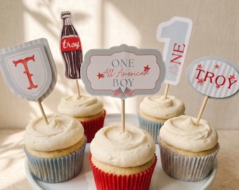 May include: Five cupcakes with white frosting in red and blue paper liners, each topped with a decorative paper topper. The toppers feature a letter T, a soda bottle labeled Troy, a sign reading ONE All American BOY, a number 1, and a sign reading TROY.