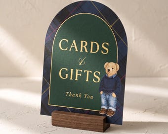 May include: An arched sign featuring a dark green center with gold text reading CARDS & GIFTS and Thank You, surrounded by a navy blue and green plaid border. A cartoon bear wearing a navy sweater and blue jeans stands on the sign, held by a wooden base.